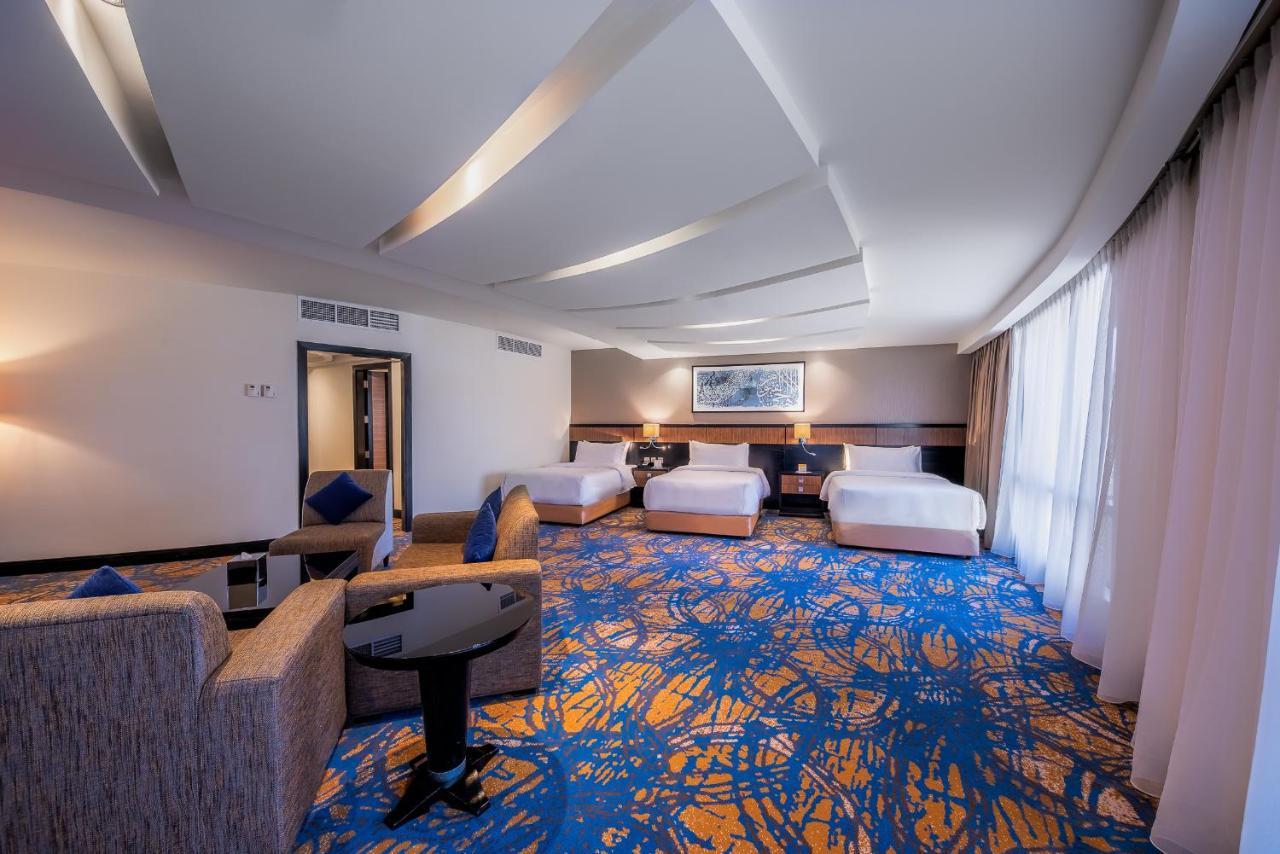 Al Ghufran Safwah Hotel Makkah Мекка Экстерьер фото The photo shows a spacious hotel room with modern decor. It features three white beds arranged neatly, a cozy seating area with a gray sofa and a black coffee table, and large windows letting in natural light. The floor has a vibrant, patterned carpe