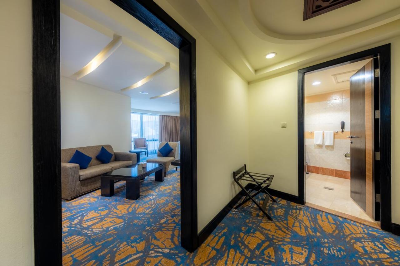 Al Ghufran Safwah Hotel Makkah Мекка Экстерьер фото The photo shows a view of a hotel room entrance. On the left side, there is a seating area with a sofa and a coffee table, decorated with blue cushions. Through a window, natural light illuminates the space. To the right, there is an open door leadin