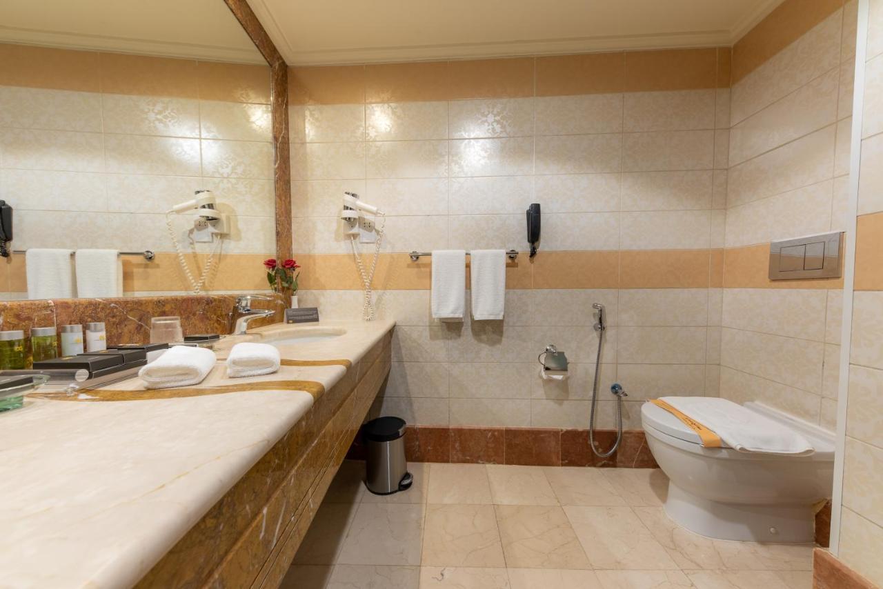 Al Ghufran Safwah Hotel Makkah Мекка Экстерьер фото The photo shows a bathroom that features a large countertop with a sink, which is made of a light-colored stone. There are several toiletries and a small vase with flowers on the countertop. Above the sink, there's a large mirror reflecting the space