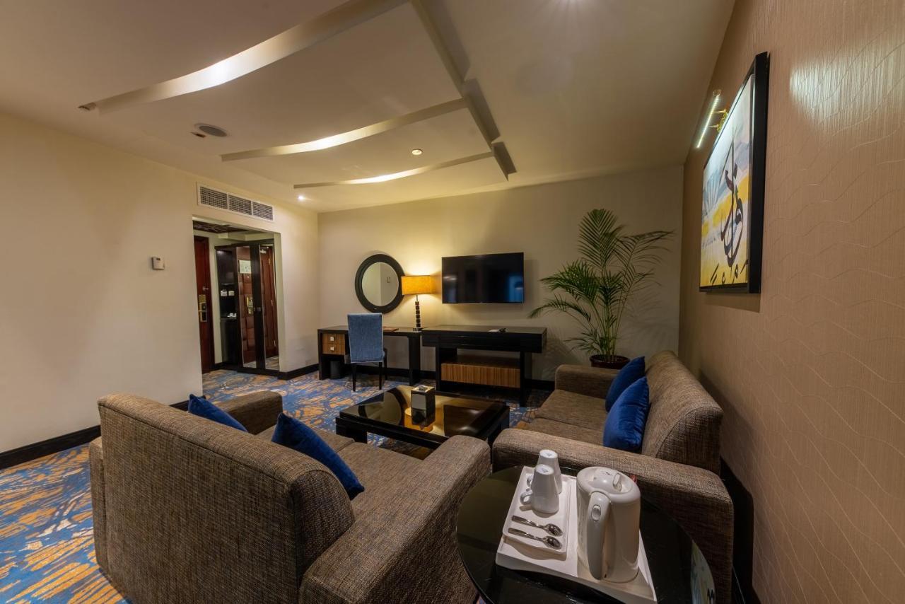 Al Ghufran Safwah Hotel Makkah Мекка Экстерьер фото The photo shows a modern living space that features a cozy seating area. There are gray sofas with blue cushions arranged around a coffee table. A small dining area or workspace with a desk and a chair is visible, along with a flat-screen television 
