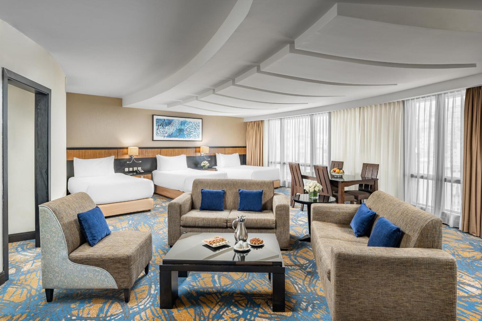 Al Ghufran Safwah Hotel Makkah Мекка Экстерьер фото The photo shows a spacious and elegantly designed hotel room. It features a cozy seating area with a coffee table surrounded by armchairs and a couch, adorned with blue cushions. In the background, there are two beds with white linens, suggesting a c