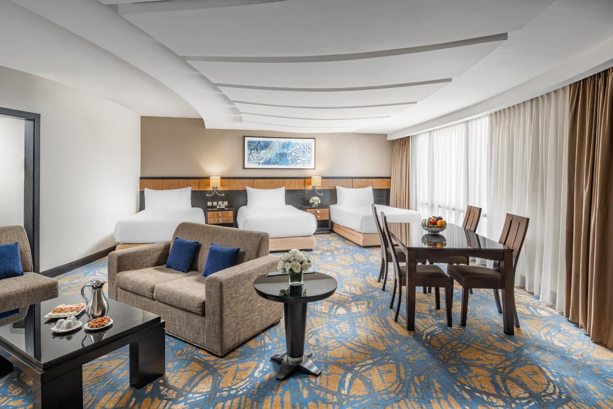 Al Ghufran Safwah Hotel Makkah Мекка Экстерьер фото The photo shows a spacious hotel room designed for comfort. It features two large beds with white bedding, a sitting area with a gray sofa and a coffee table, and a dining area with a wooden table and chairs. There is a decorative vase on the table a